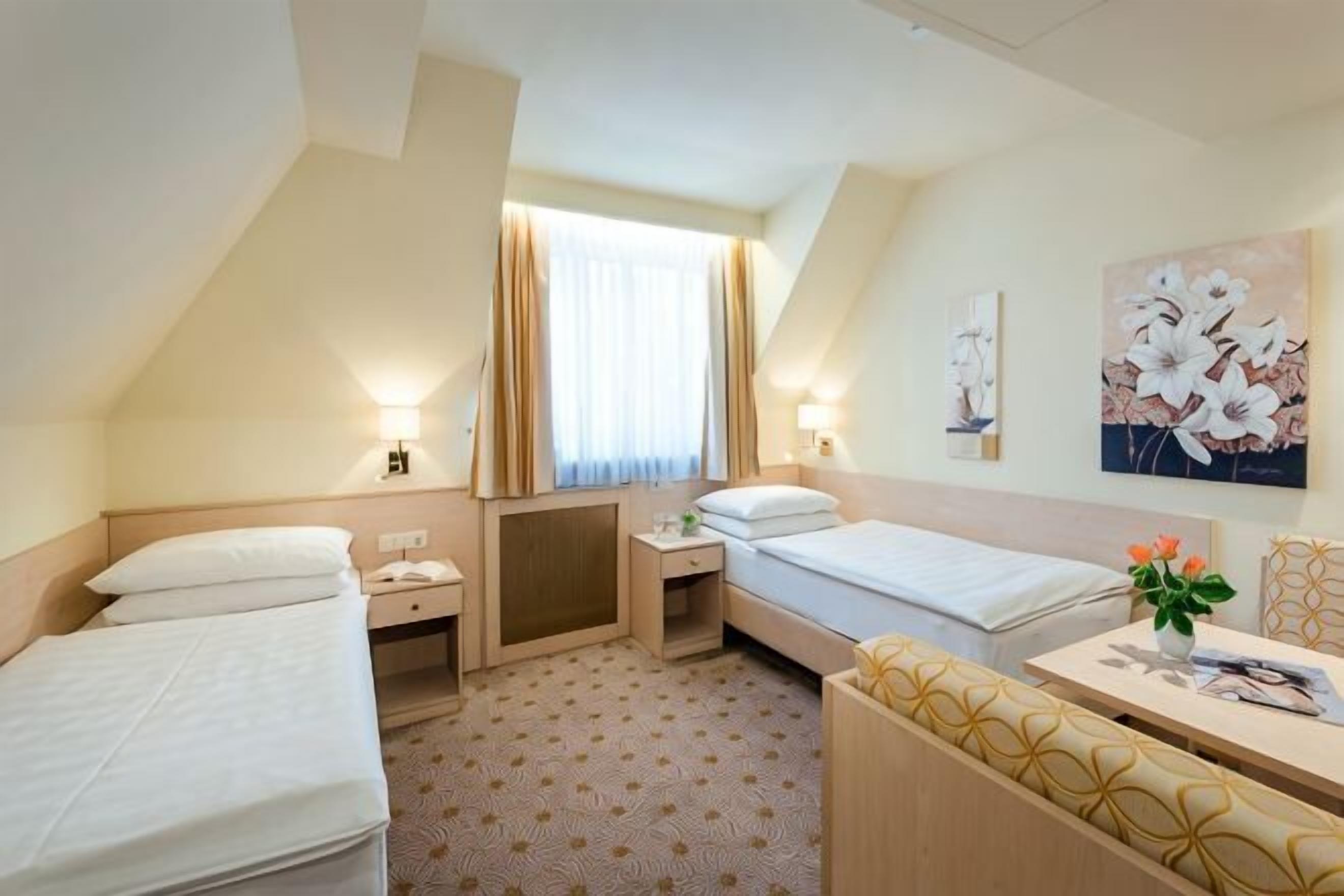 Une chambre d’hôtel avec deux lits, un bureau, une chaise, une fenêtre avec des rideaux et des œuvres d’art accrochées aux murs.