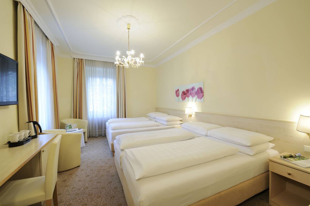 A hotel room with three beds, a desk with a chair, a TV, and a chandelier.