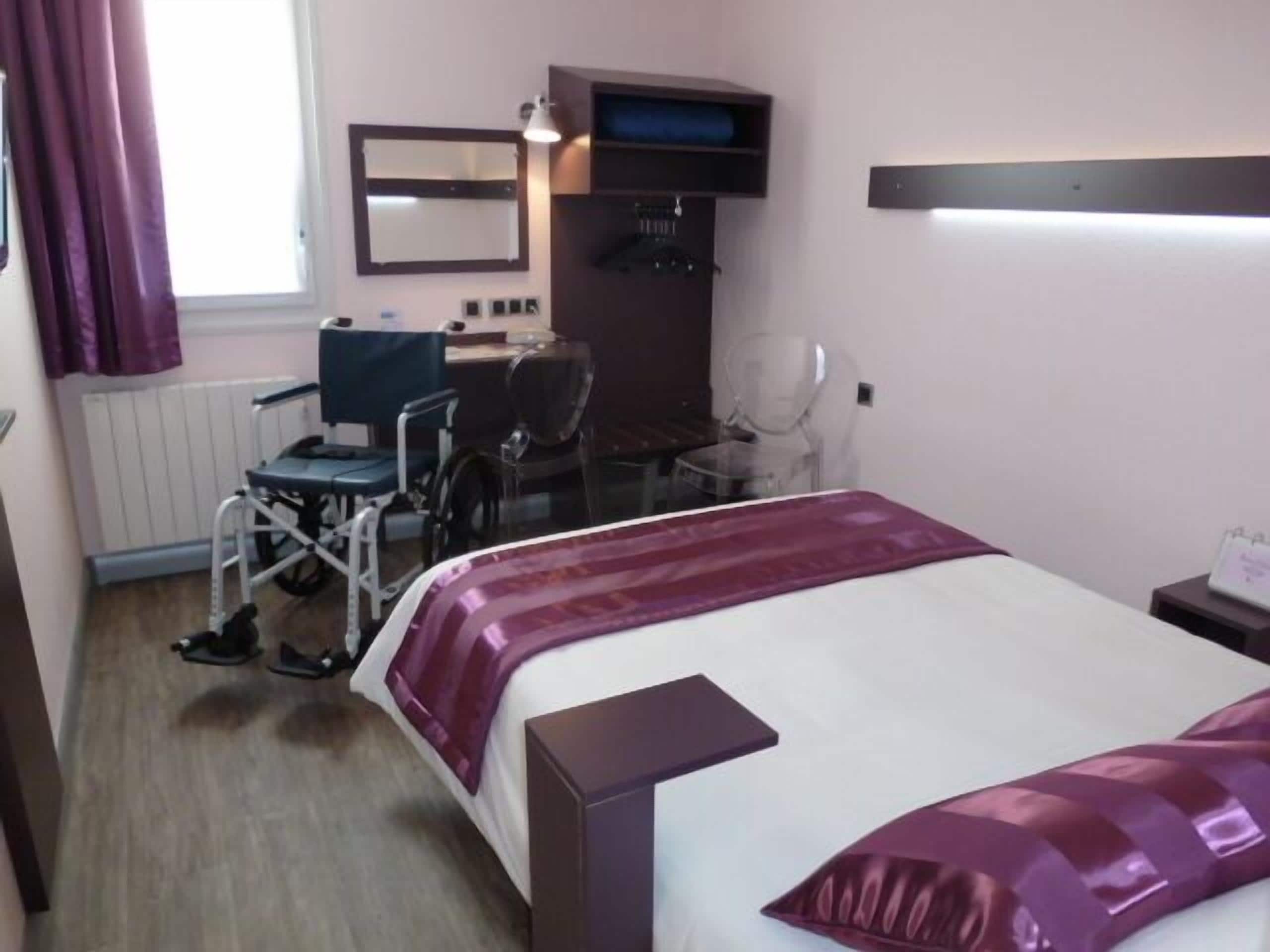 A hotel room with a bed, a desk, a chair, a wheelchair, and a window with curtains.