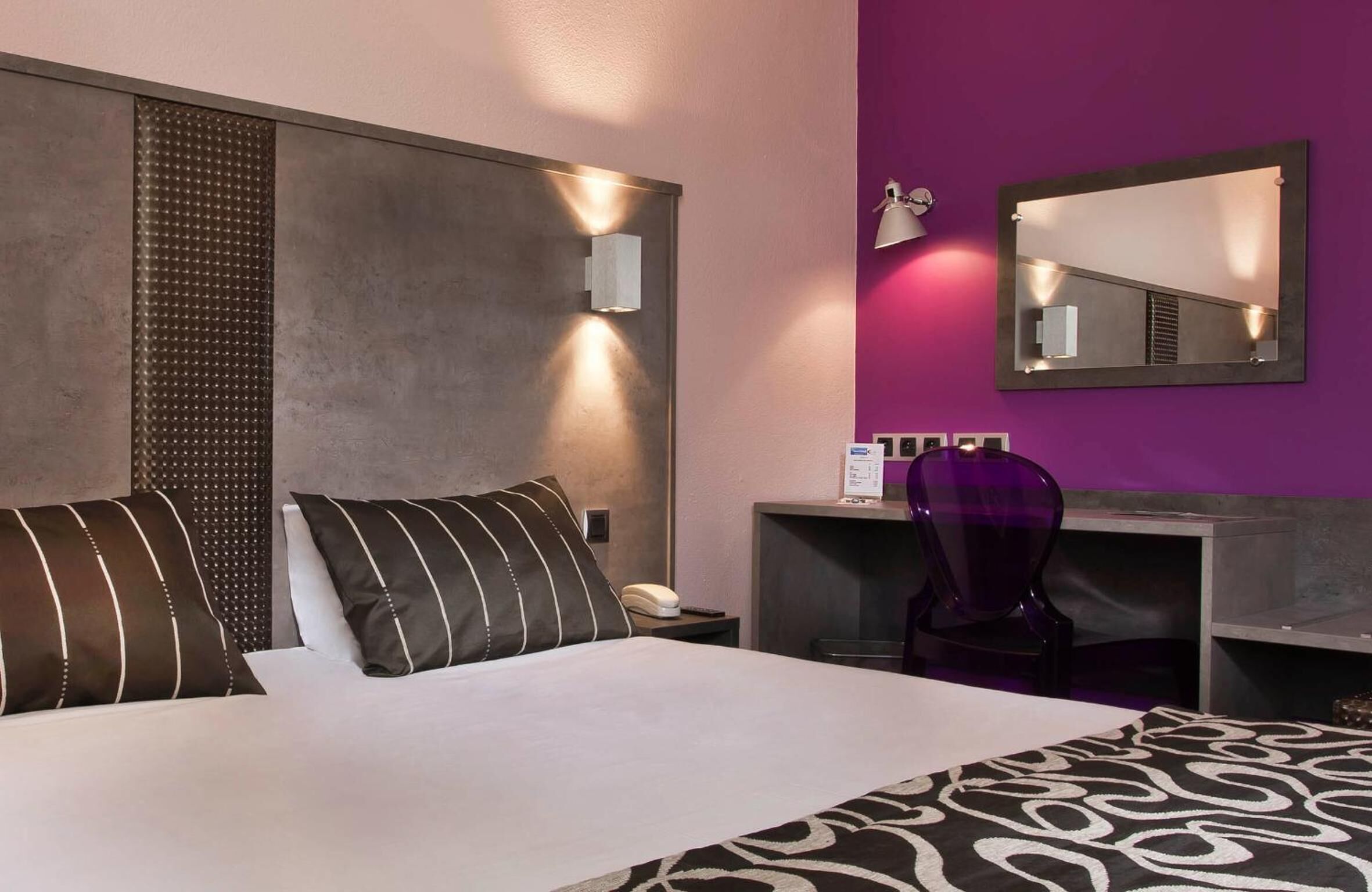 A hotel room with a large bed, a desk with a chair, a mirror, and a wall-mounted phone.