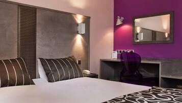 A hotel room with a large bed, a desk with a chair, a mirror, and a wall-mounted phone.