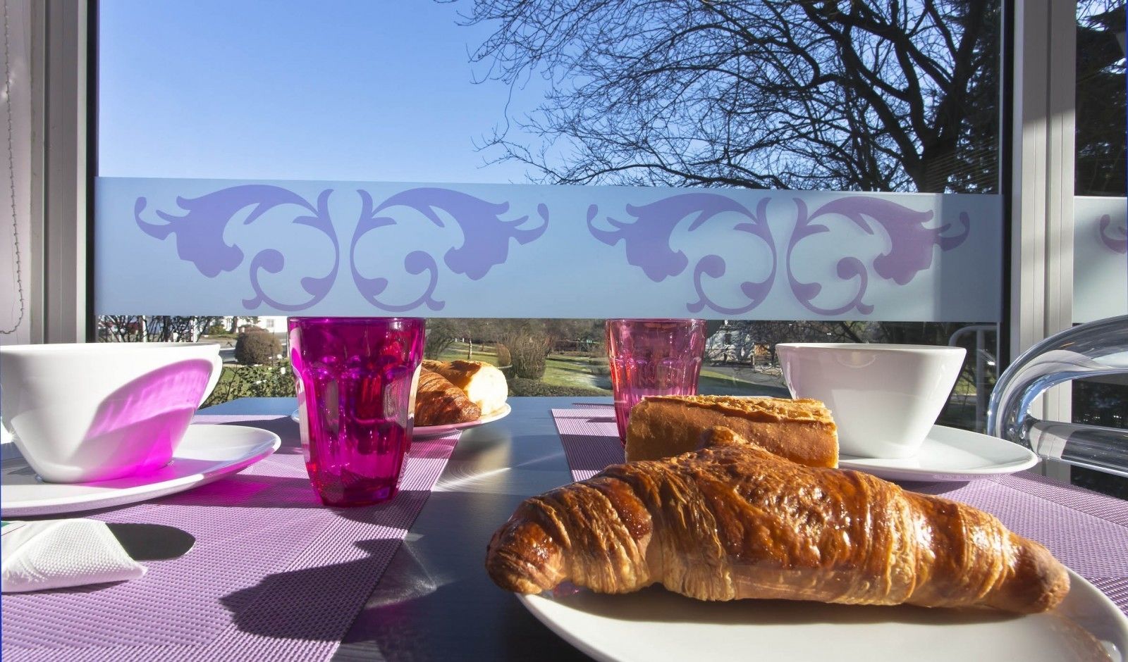 Una mesa con un croissant, dos tazas y un plato de croissants, frente a una ventana con vistas a árboles y un cielo despejado.