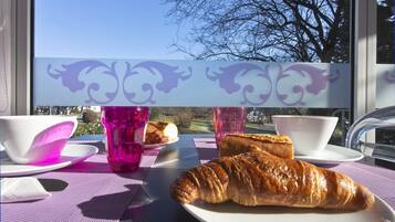 Una mesa con un croissant, dos tazas y un plato de croissants, frente a una ventana con vistas a árboles y un cielo despejado.