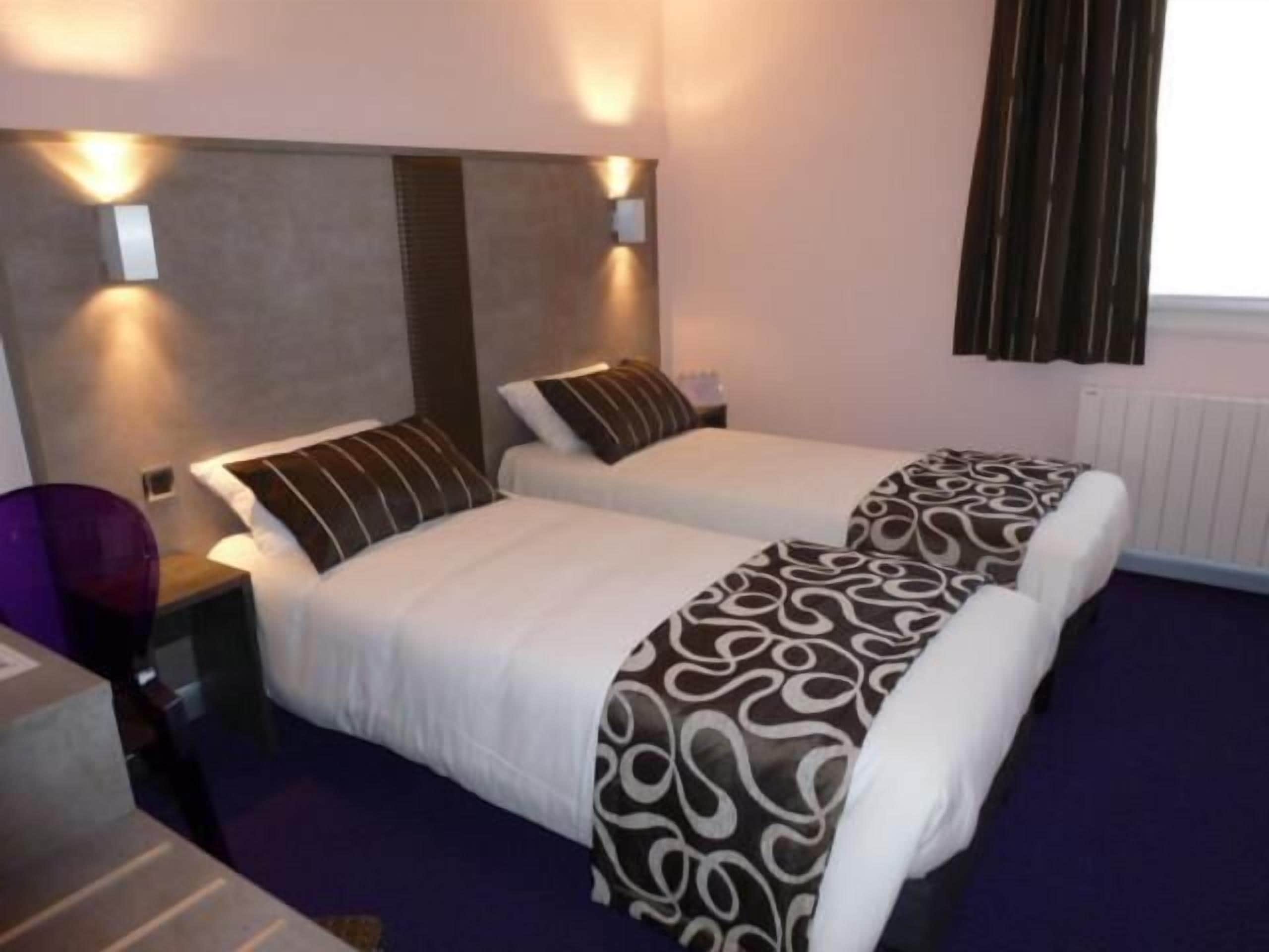 A hotel room with two beds, each with a pillow and a blanket. There are wall-mounted lights above the beds and a window with curtains.