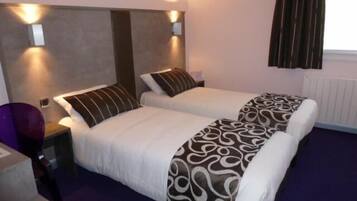 A hotel room with two beds, each with a pillow and a blanket. There are wall-mounted lights above the beds and a window with curtains.