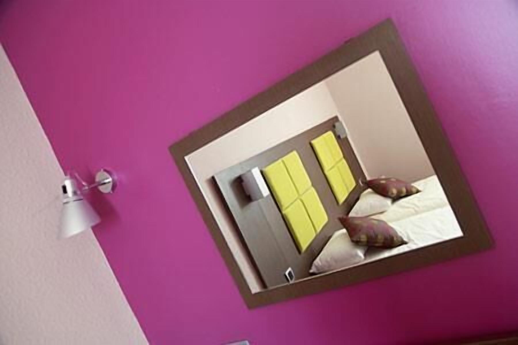 A hotel room with a purple wall, a mirror reflecting a bed with pillows, and a white lamp on the wall.