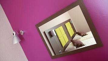 A hotel room with a purple wall, a mirror reflecting a bed with pillows, and a white lamp on the wall.