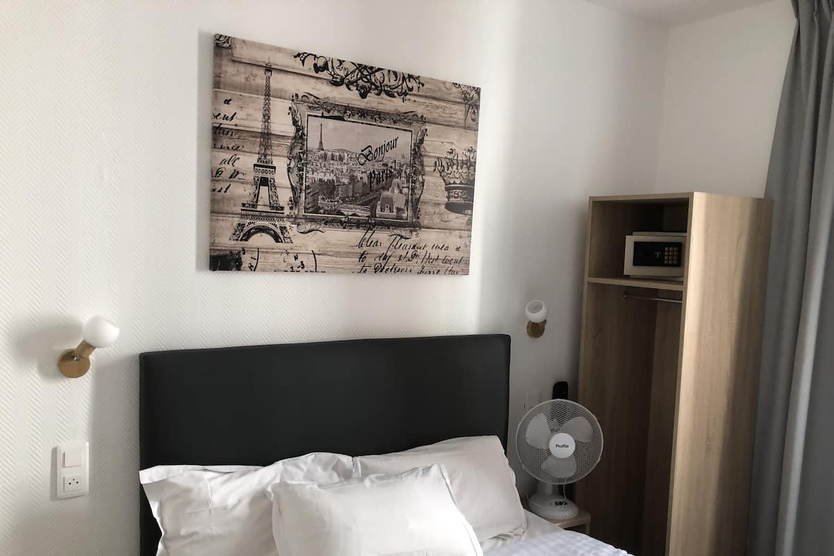 A neatly made bed with white linens and pillows, a wall-mounted picture of Paris, a bedside table with a lamp, and a small fan.