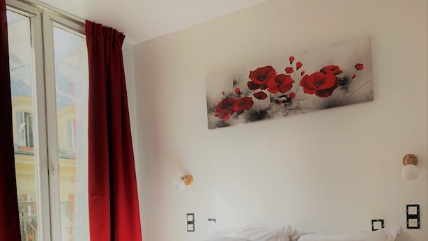 Una cama bien hecha con sábanas blancas, una cortina roja entreabierta y un cuadro de flores rojas en la pared.