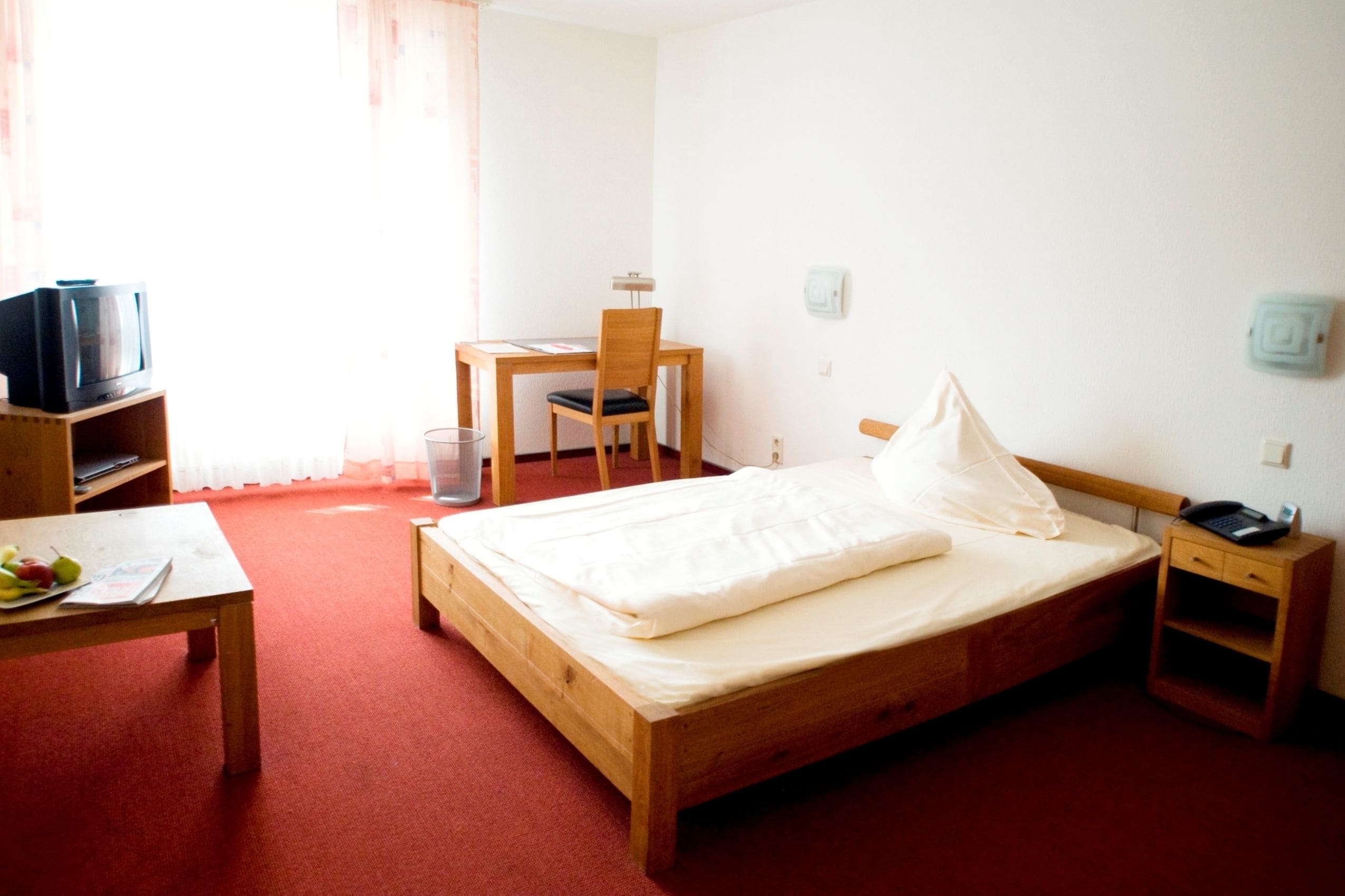 Une chambre d’hôtel avec un lit en bois, un bureau avec une chaise, un téléviseur posé sur une étagère et une petite table de chevet.