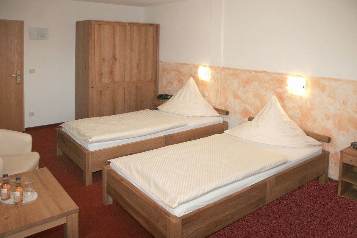 Quarto com duas camas de madeira, um guarda-roupa de madeira, uma mesinha com garrafas e um tapete vermelho.