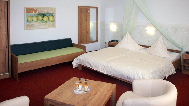 Une chambre d’hôtel avec un lit, un canapé vert, une table en bois avec des bouteilles, une chaise blanche et un tableau au mur.