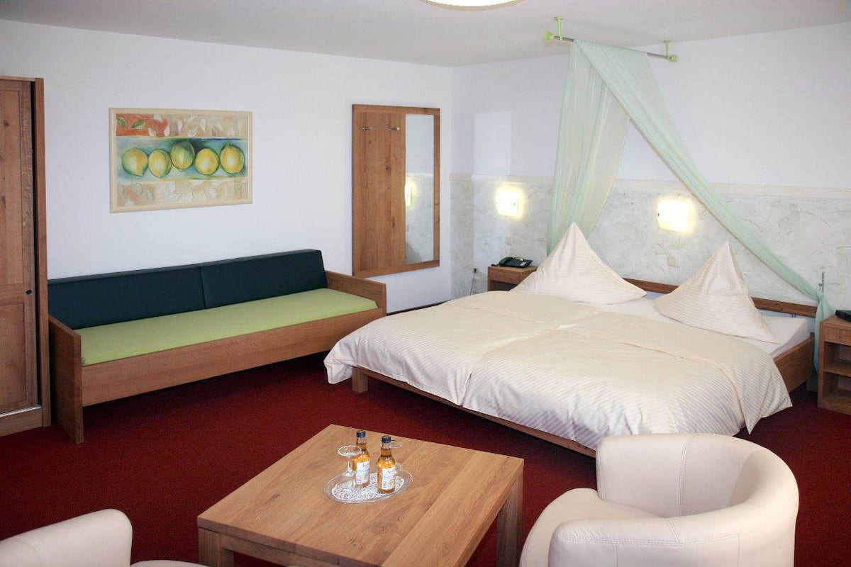 Quarto de hotel com cama, sofá verde, mesa de madeira com garrafas, cadeira branca e um quadro na parede.