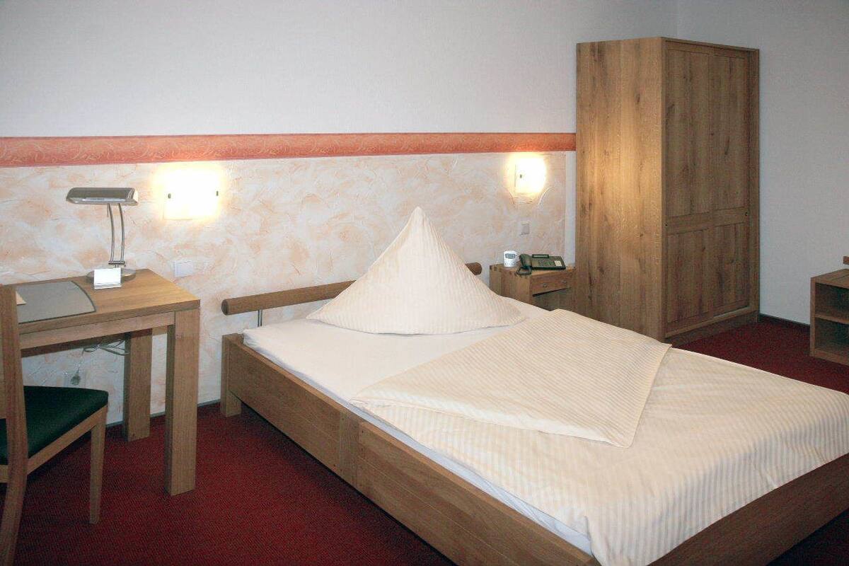Uma cama de solteiro com estrutura de madeira, uma mesa de cabeceira com abajur, um guarda-roupa de madeira e uma pequena escrivaninha com cadeira.