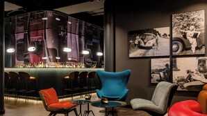 Una zona de bar con diseño moderno, que incluye una barra con botellas, un área de asientos con sillas y cuadros de autos antiguos colgados en la pared.