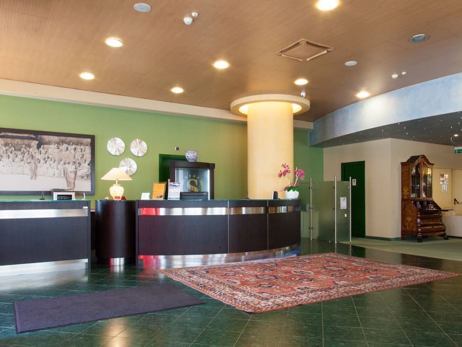 A reception area with a reception desk, a large wall clock, a decorative vase with flowers, and a patterned rug on the floor.