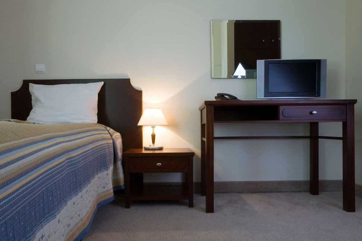 A hotel room with a bed, a nightstand with a lamp, a desk with a computer, and a mirror on the wall.