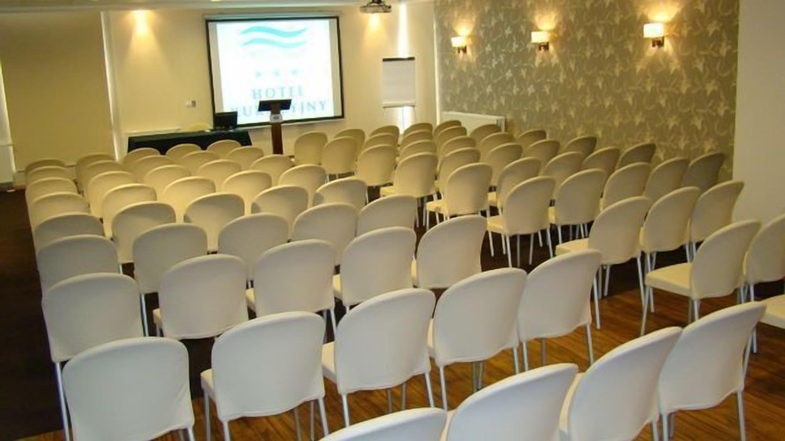 Una sala de conferències amb files de cadires blanques, una pantalla de projecció i un atril.