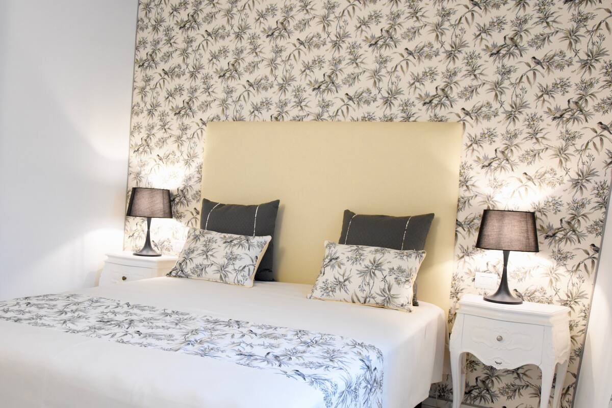 A bedroom with a large bed, floral wallpaper, two nightstands with lamps, and matching pillows.