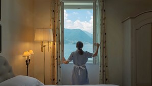 Una habitación con cama, lámpara, cortinas y ventana con vista a una montaña y agua.