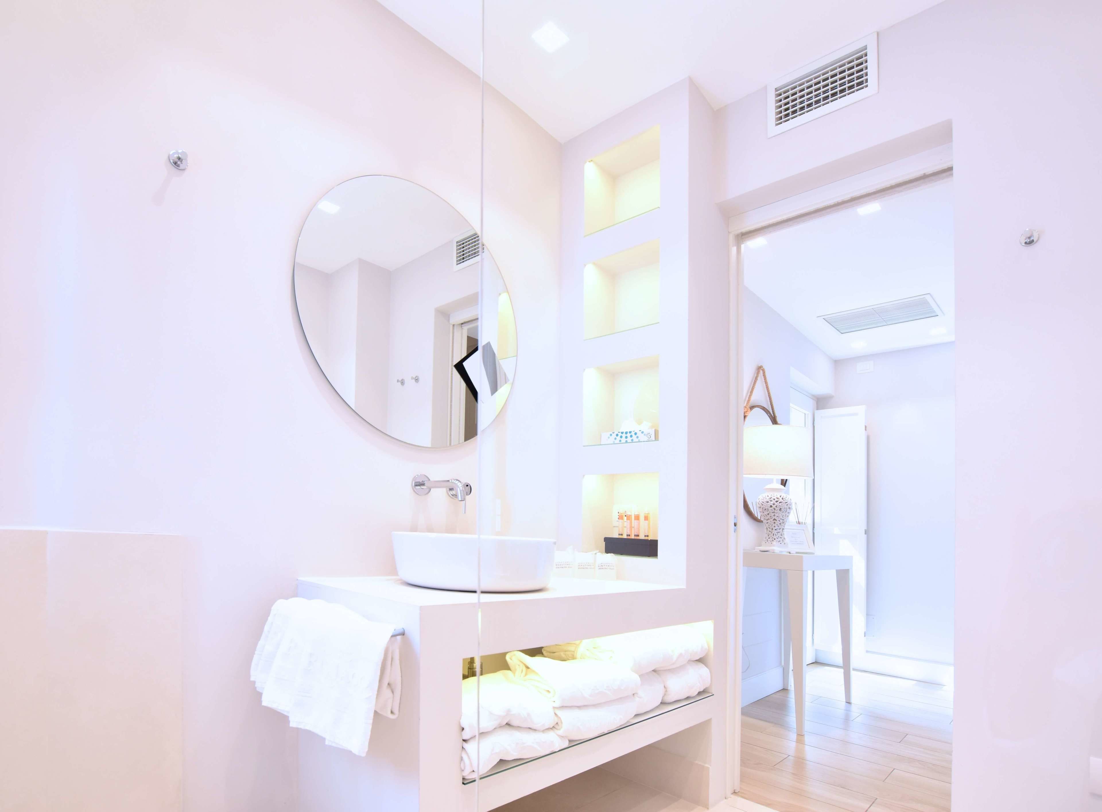 Un baño moderno con un espejo redondo, tocador blanco y repisas con artículos de aseo.