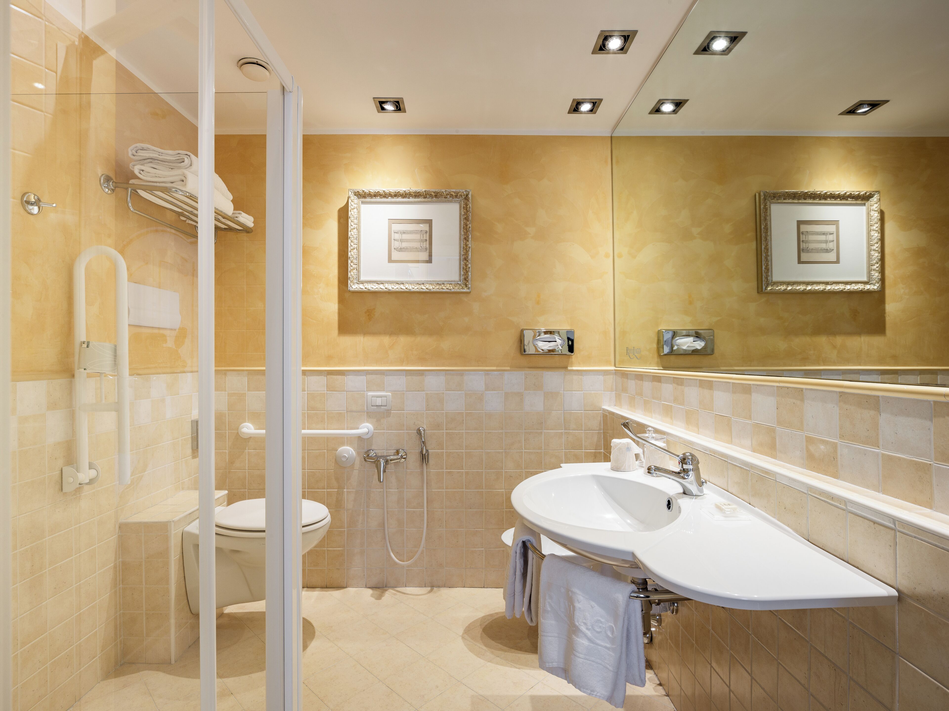 Un baño con cabina de ducha de vidrio, inodoro, lavamanos con toallero y espejo con fotografías enmarcadas.