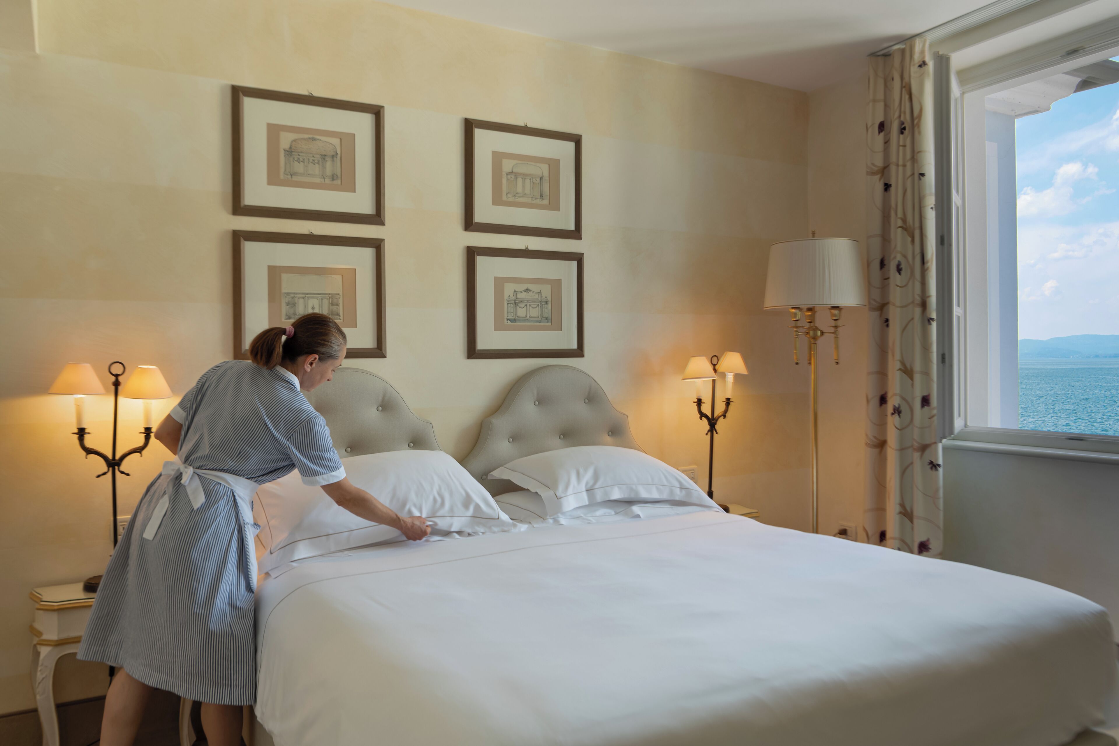 Una cama bien tendida en una habitación de hotel con cuadros en la pared, una lámpara y vista al mar.