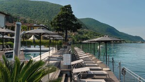 Un hotel junto al lago con una zona de lounge junto a la piscina, sombrillas y vistas al lago y las colinas.