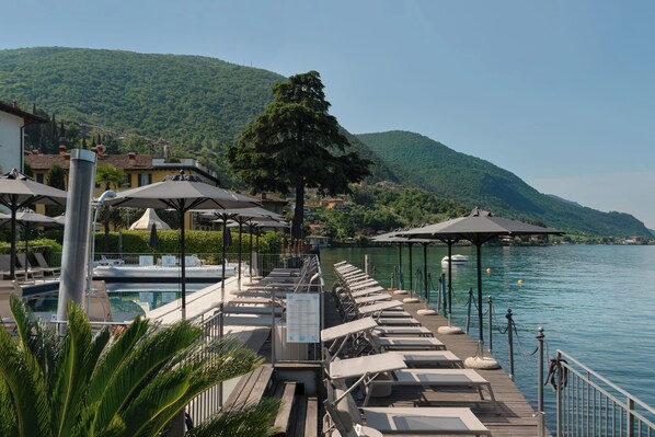 A lakeside hotel with a poolside lounge area, umbrellas, and a view of the lake and hills.
