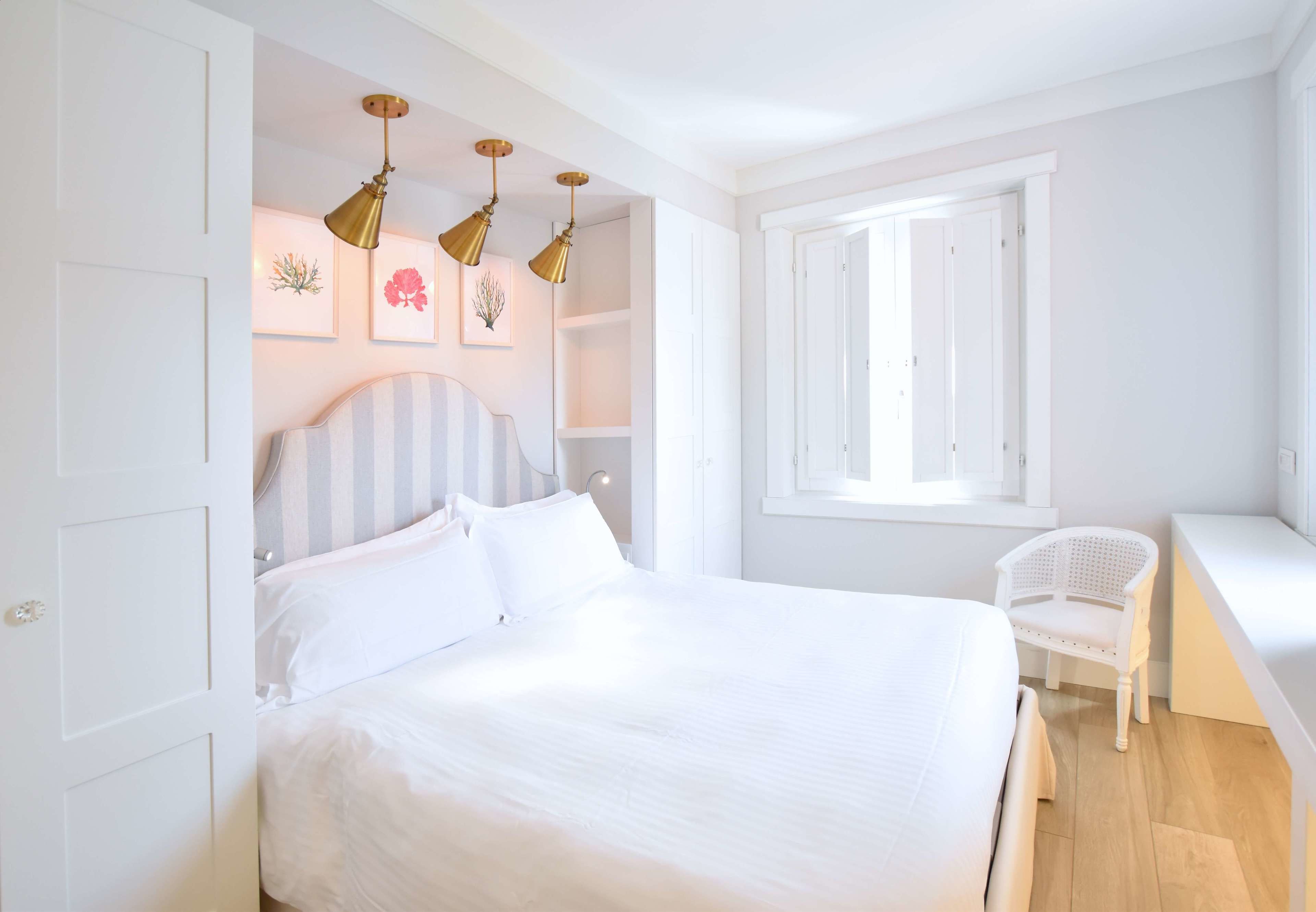 A bedroom with a large bed, a white chair, and a window with open shutters.