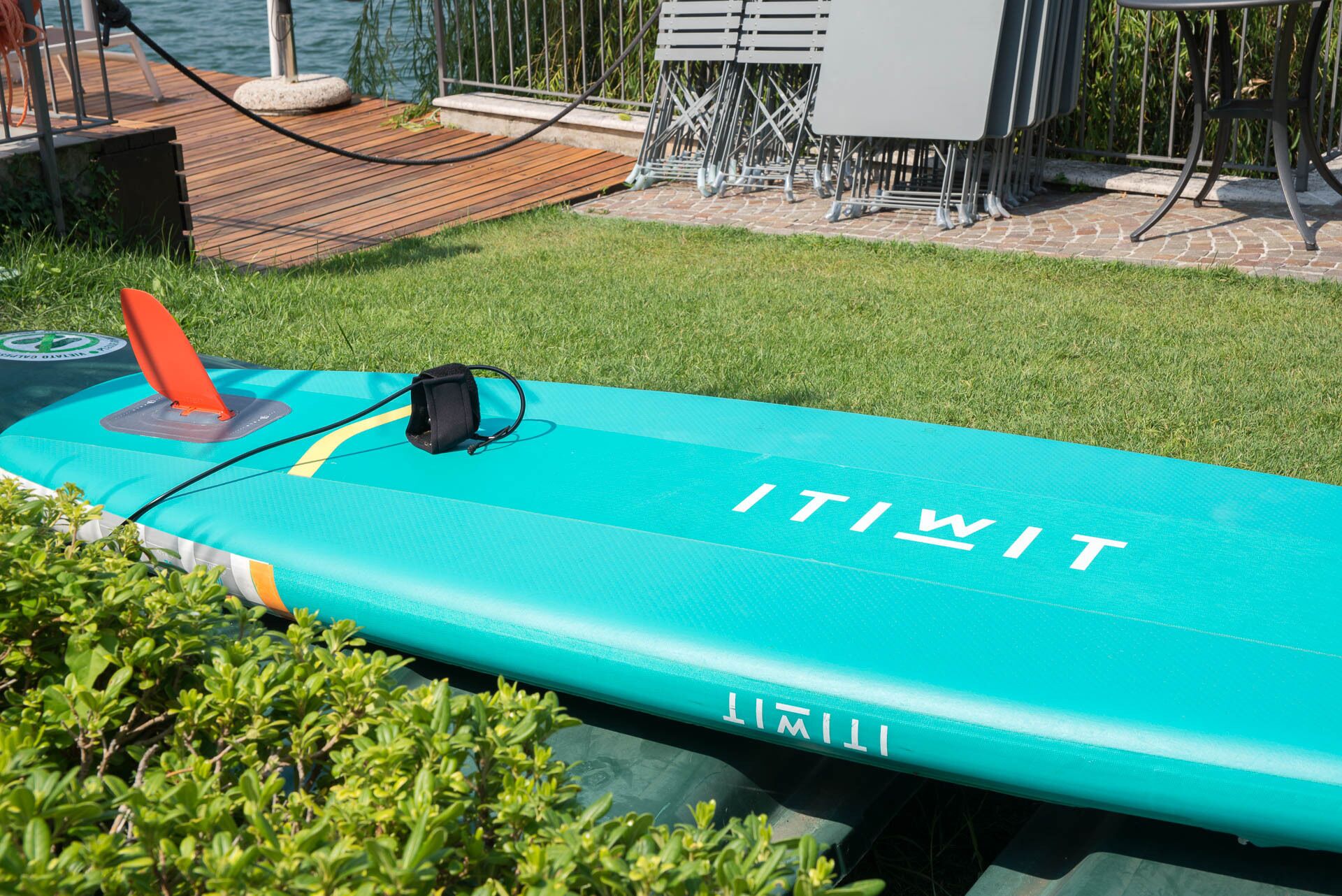 Uma prancha de surf insuflável azul com a inscrição "ITIWIT", colocada numa área gramada perto de um cais e área de estar ao ar livre.