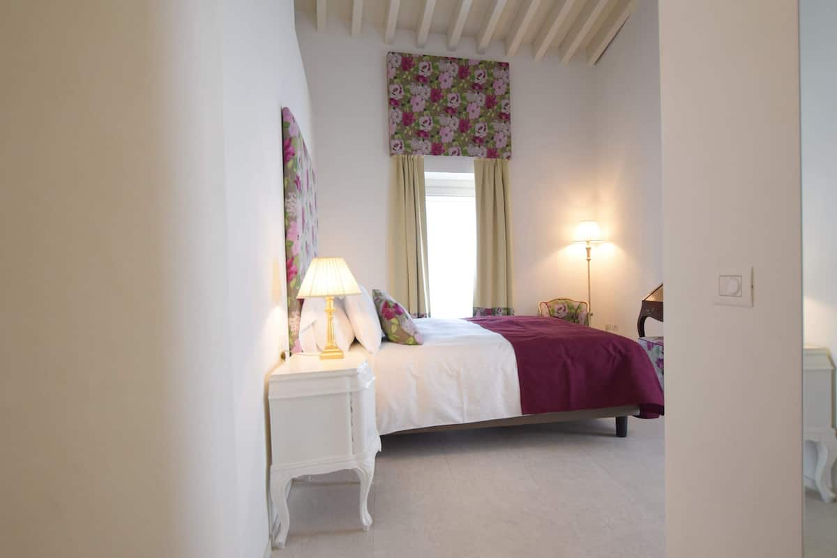 A bedroom with a bed, a nightstand with a lamp, a window with curtains, and a wall-mounted light fixture.
