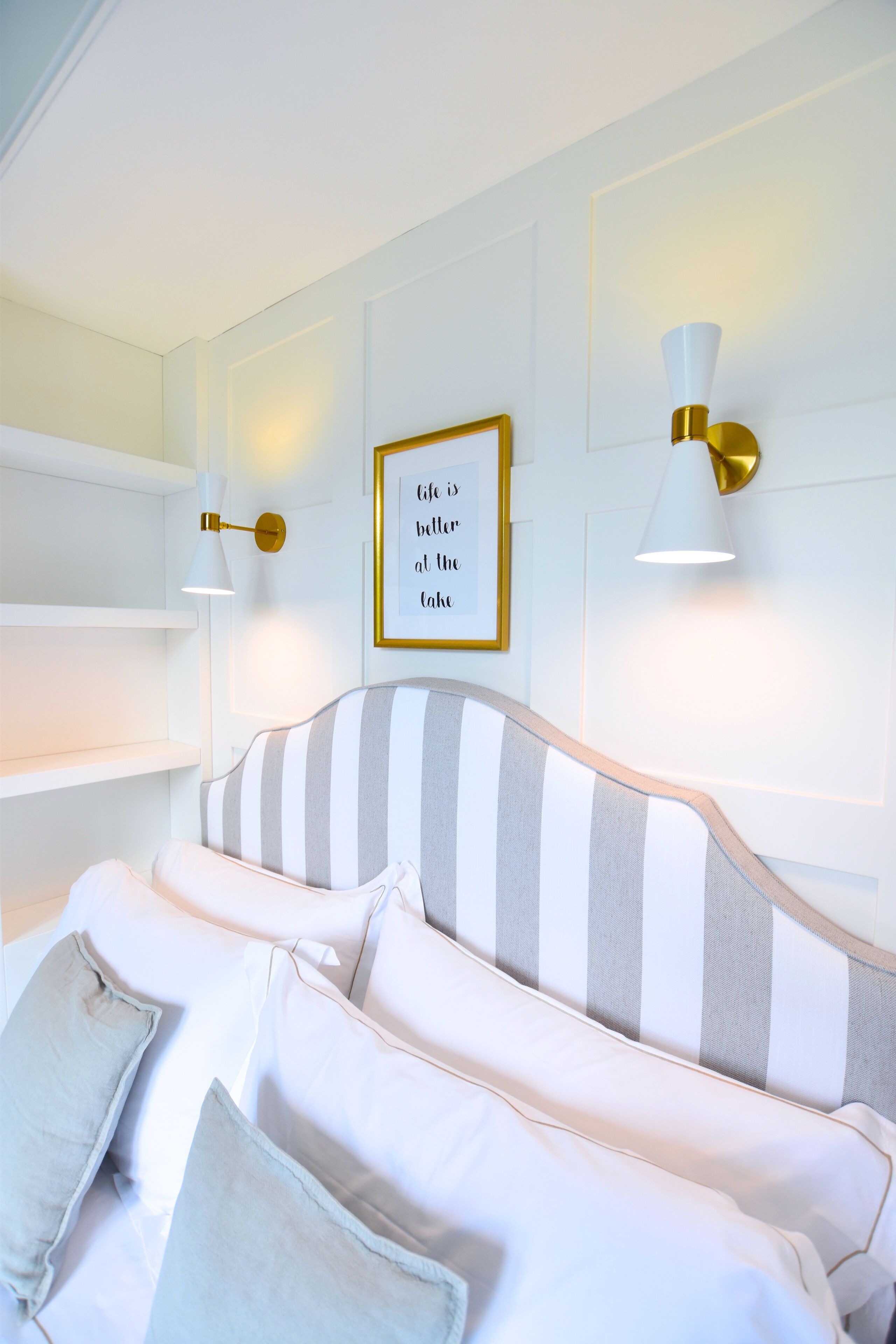 Un dormitorio ordenado con un cabecero a rayas, ropa de cama blanca y dos lámparas fijadas a la pared.