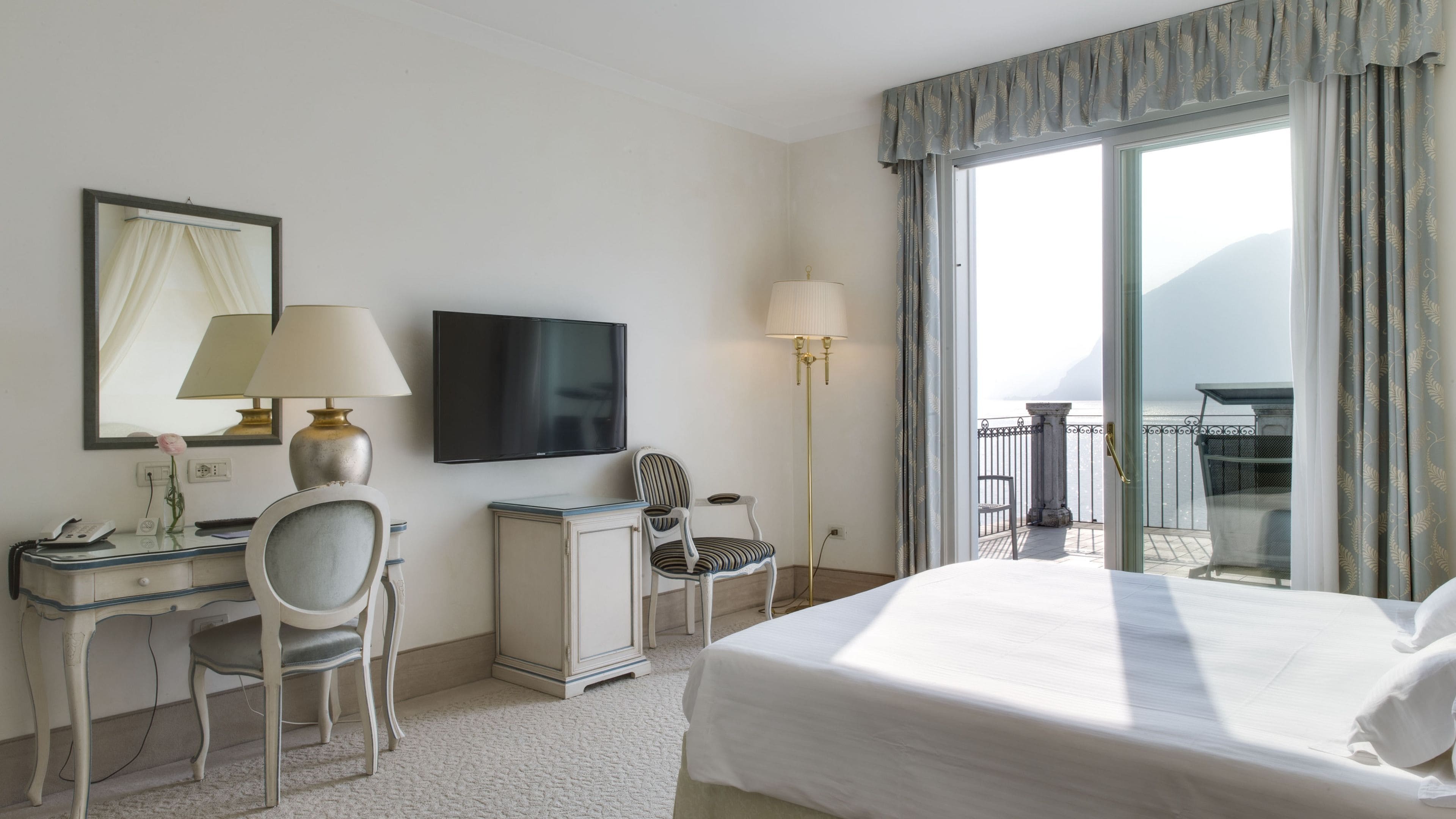 A hotel room with a bed, desk, chair, TV, and a balcony with a view.