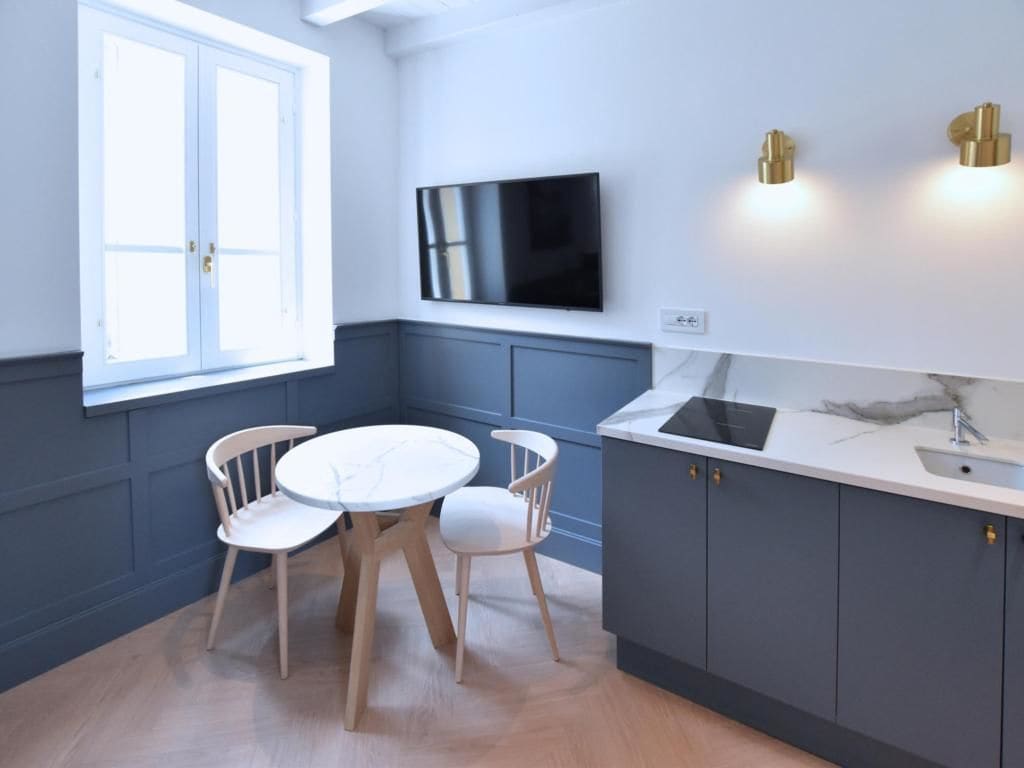 A modern kitchen with a dining table and chairs, a flat-screen TV mounted on the wall, and a window with a door.