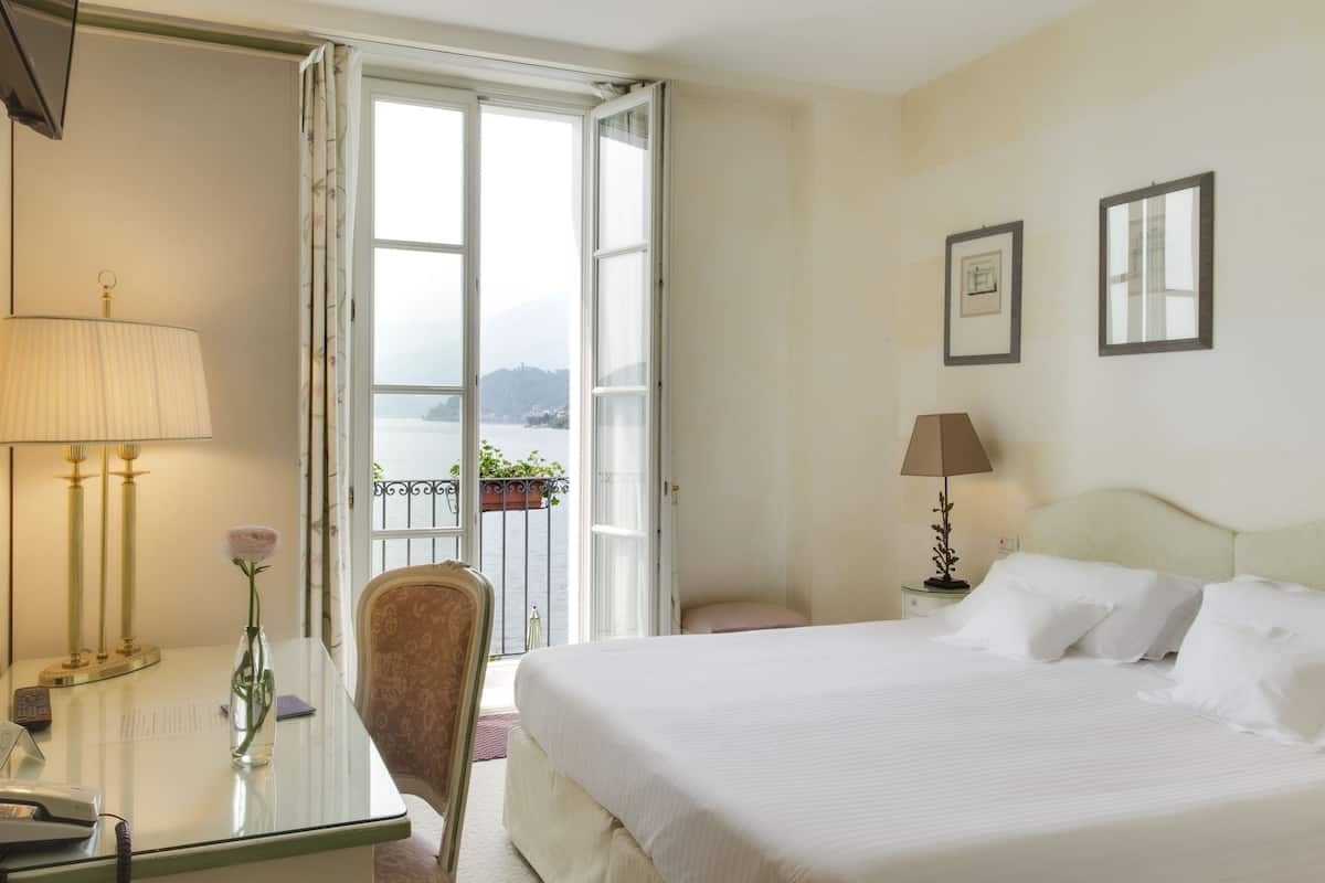 A hotel room with a bed, a desk with a lamp, a chair, a window with a view of a body of water, and a balcony with a plant.