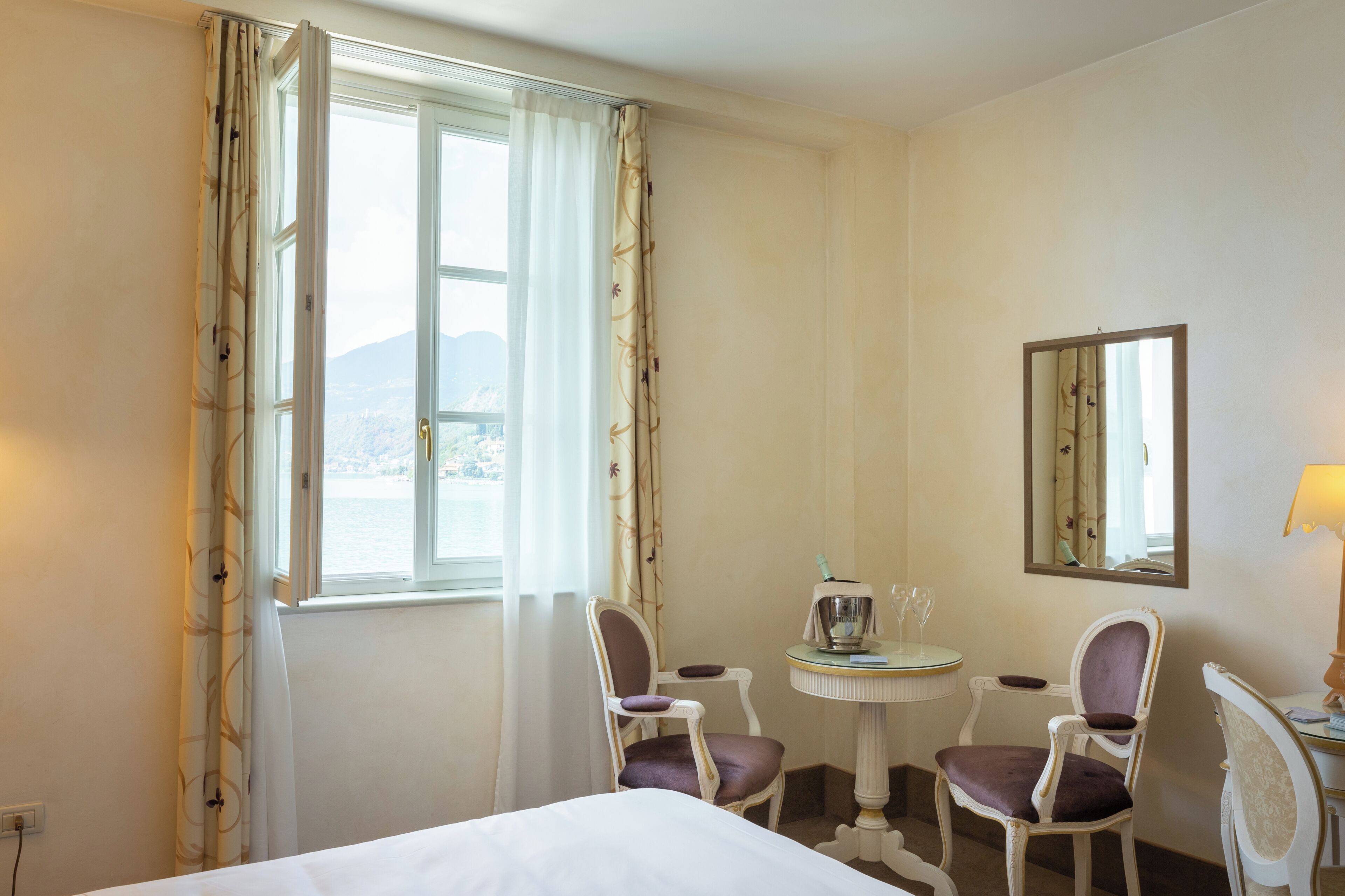 A hotel room with a bed, two chairs, a small table with a lamp, a mirror, and a window with curtains.