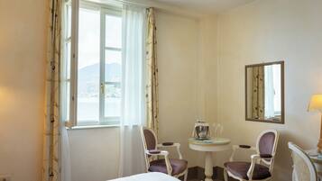 A hotel room with a bed, two chairs, a small table with a lamp, a mirror, and a window with curtains.
