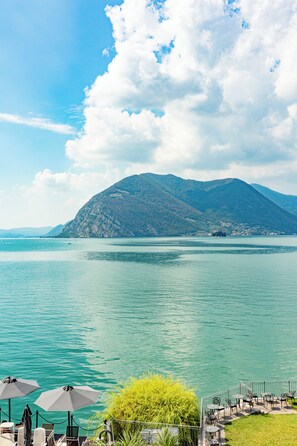A lakeside view with mountains, outdoor seating, and umbrellas.