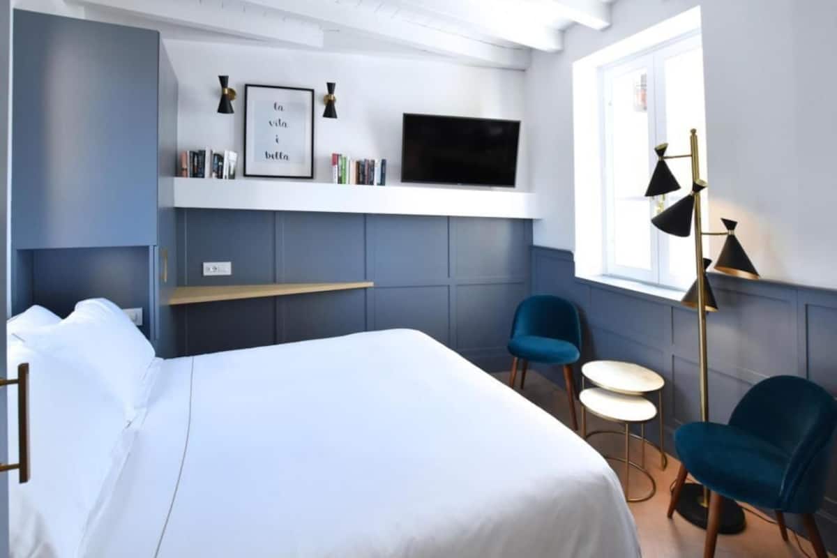 A modern hotel room with a bed, a TV mounted on the wall, a desk, and two chairs.