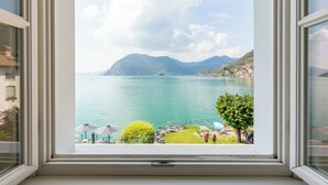 Vista desde una ventana sobre un lago con montañas al fondo, un área verde con asientos al aire libre y un árbol.