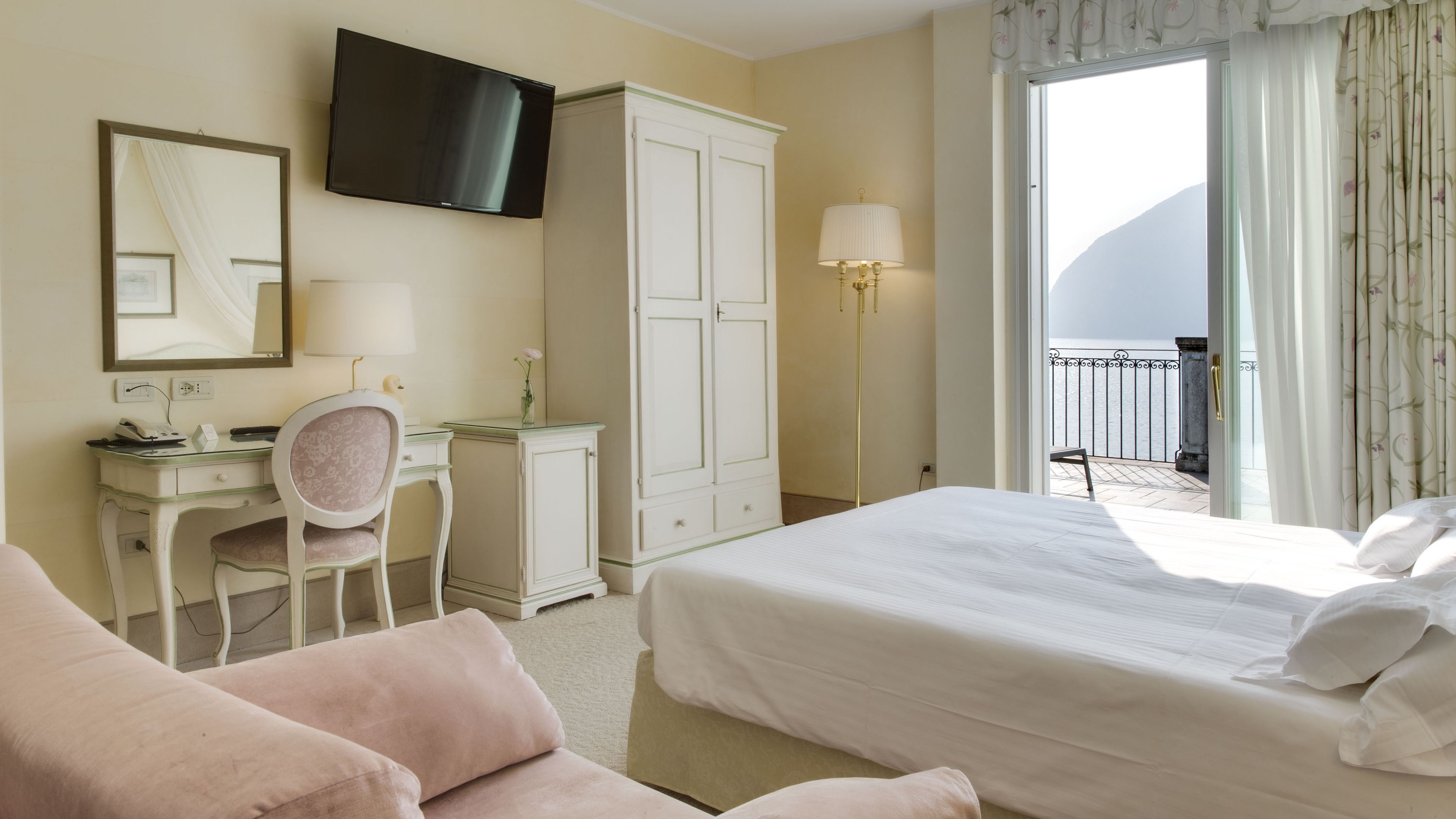 Habitación de hotel con cama, escritorio y silla, armario, televisión y balcón con cortinas.