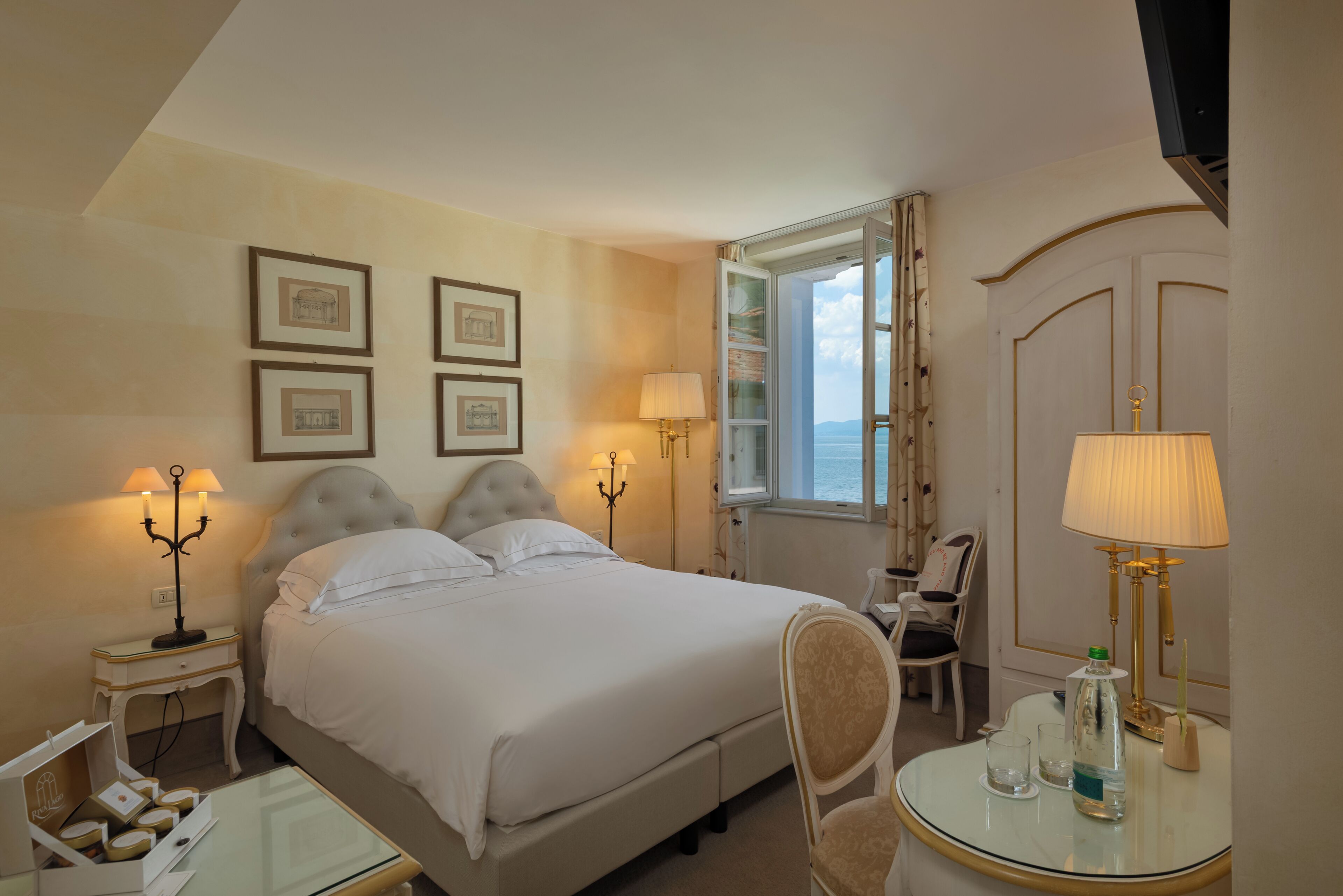 A hotel room with a bed, bedside lamps, a chair, a table with a bottle and glasses, and framed pictures on the wall.