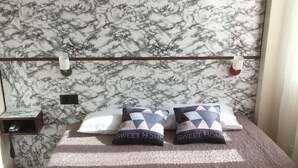 Una cama bien hecha con un cabecero estampado con diseño de mármol, dos almohadas con el texto 'SWEET HOME' y dos toallas dobladas sobre la cama.