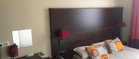 Habitación de hotel con cabecera de madera, dos lámparas de noche, cama con almohadas estampadas, mesita de noche con un teléfono y un cartel de 'No Fumar', y una obra de arte colgada en la pared sobre la cama.
