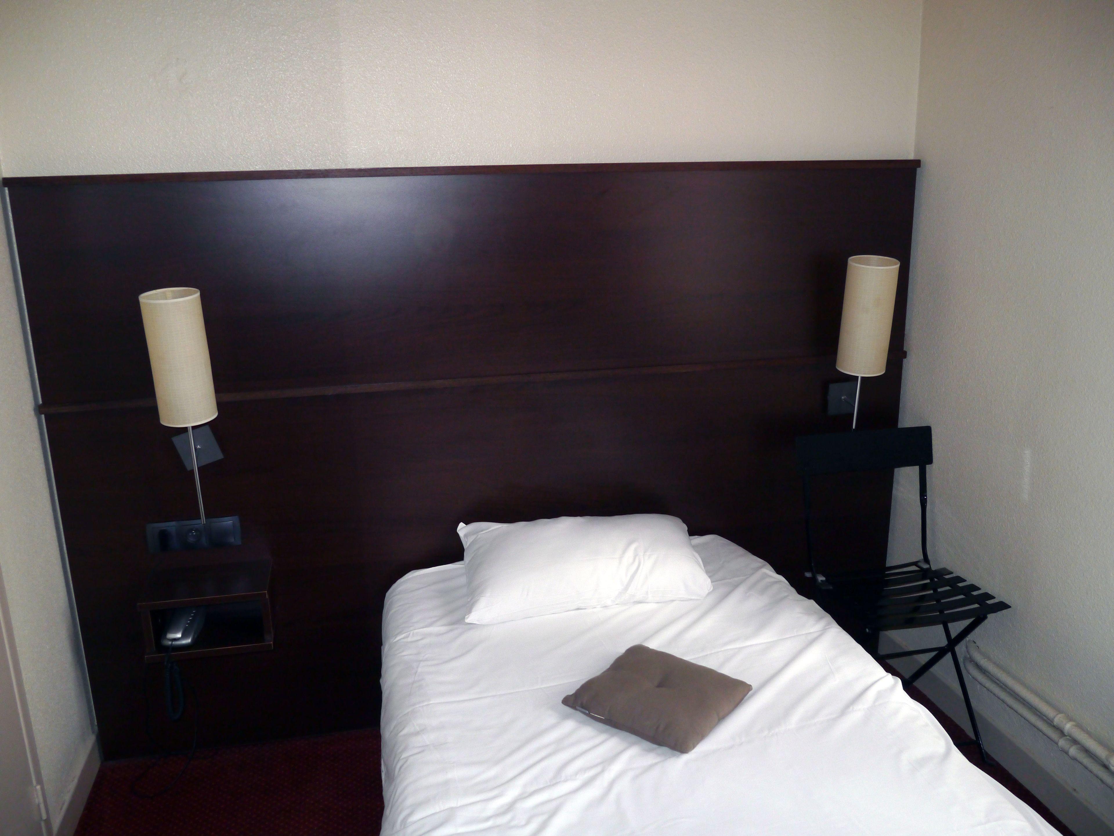 Habitación de hotel con una cama individual, ropa de cama blanca, un almohadón marrón, dos lámparas de noche y un pequeño estante con un control remoto.