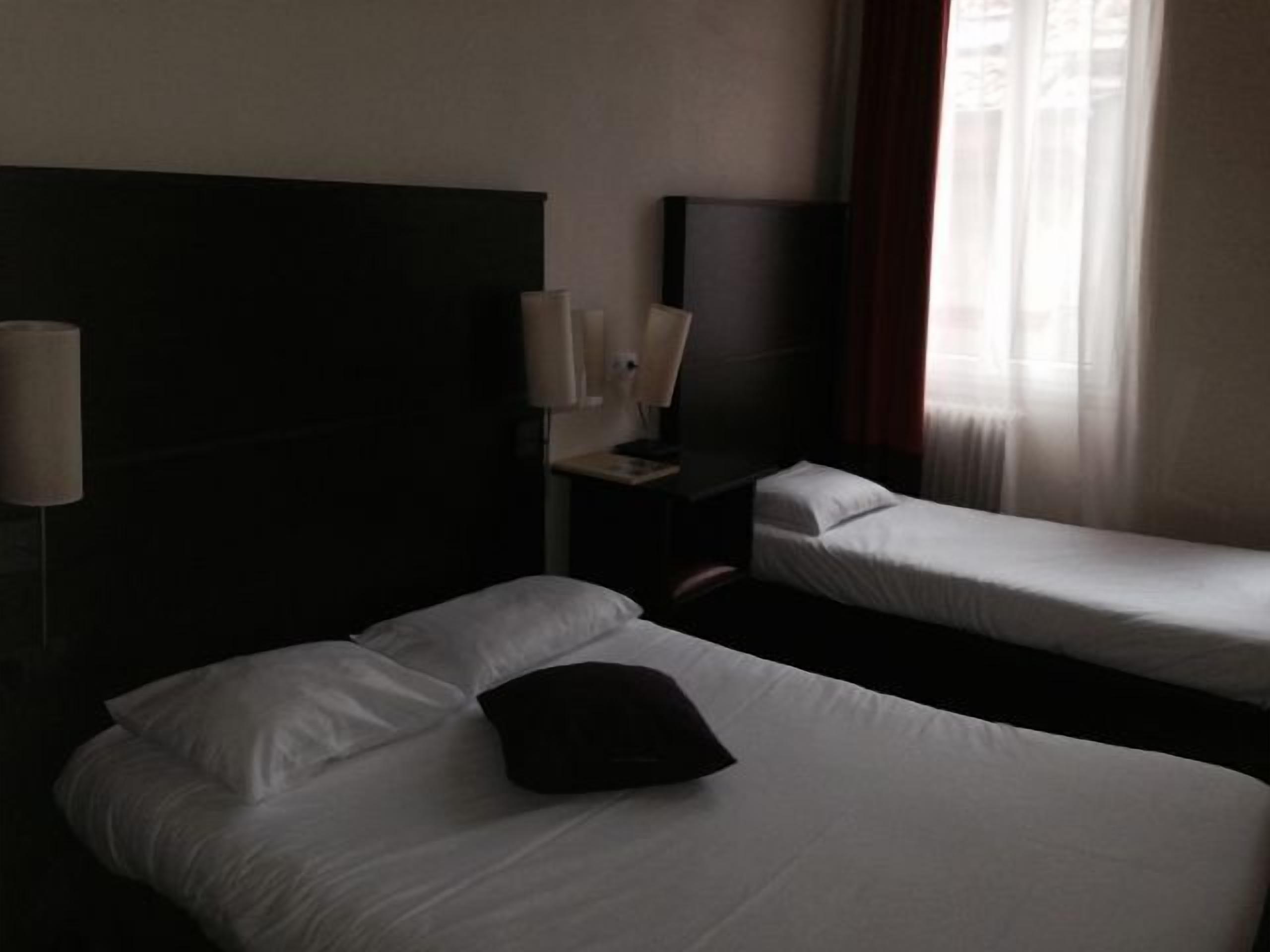 Habitación de hotel con dos camas individuales, una mesita de noche con lámpara y una ventana con cortinas.