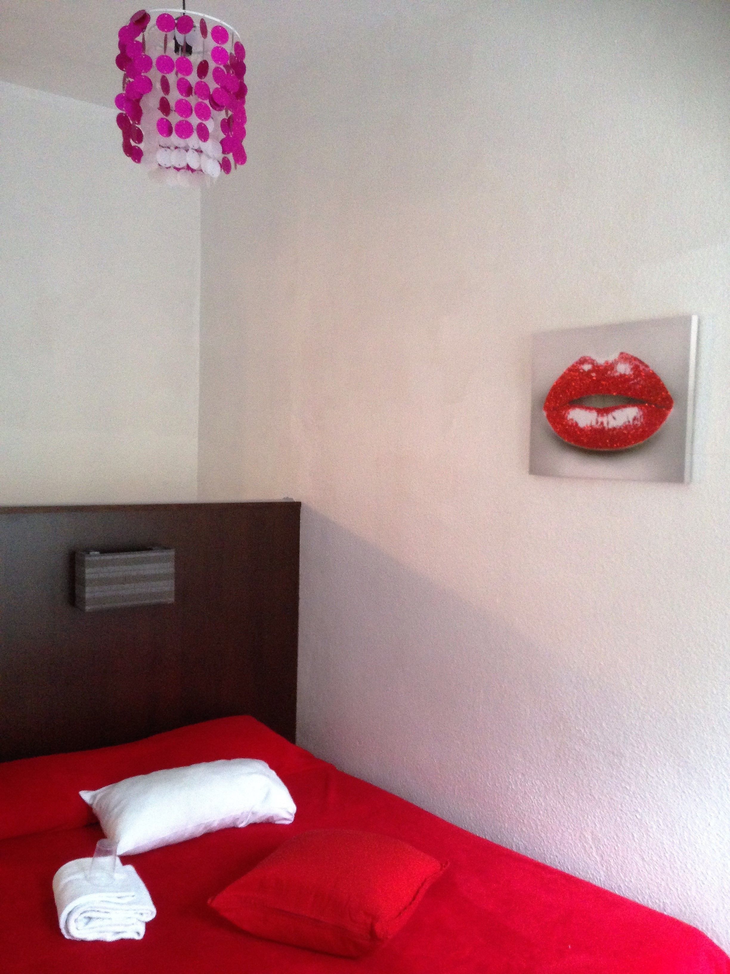 A hotel room with a red bed, white pillows, a red throw blanket, a white towel, a headboard, and a wall-mounted artwork of red lips.