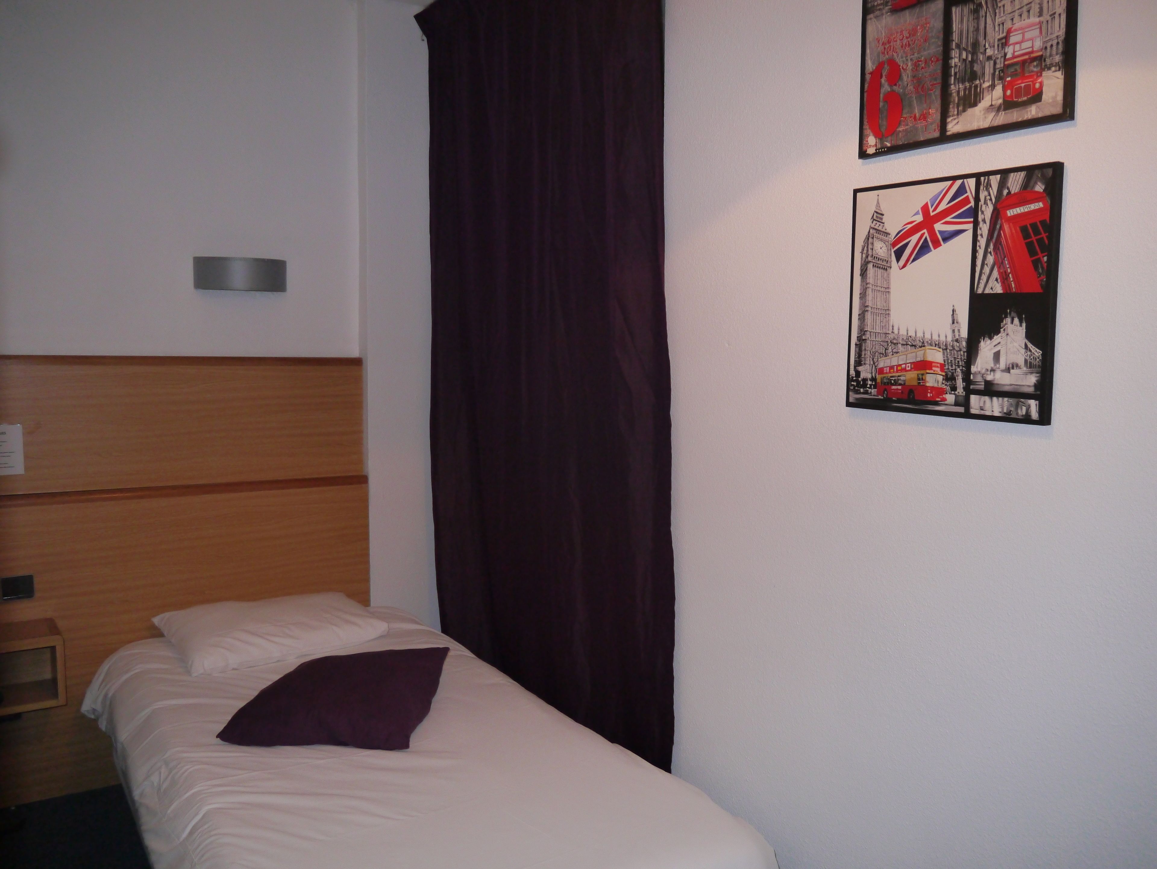 Ein Einzelbett mit weißer Matratze und einem violetten Kissen, ein Holz-Kopfbrett, ein dunkler Vorhang und zwei gerahmte Bilder von britischen Sehenswürdigkeiten an der Wand.
