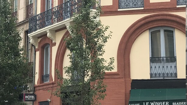 Un hotel con fachada clásica, que presenta un balcón con barandillas ornamentales y un amplio ventanal de arco.